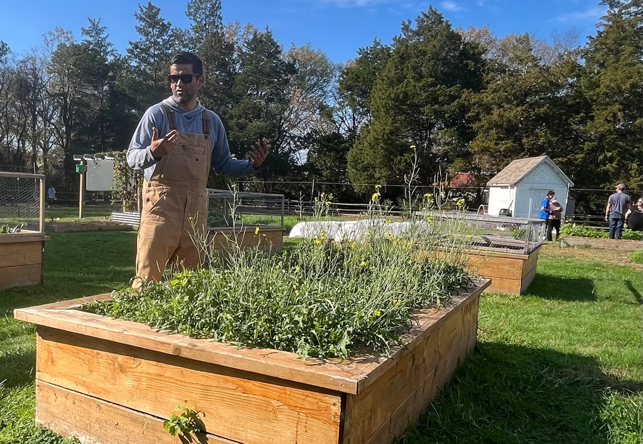A man stands next to a raise garden bed, gesturing as he speaks. Other raised garden beds can be seen in the background.